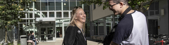 Två glada studenter, en tjej med ljust hår och en kille med mörkt hår och glasögon som parkerar sin cykel utanför campuset.