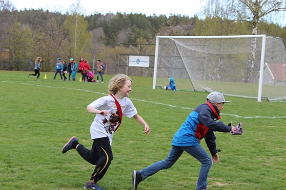 Barn som jagar varandra framför ett fotbollsmål på en fotbollsplan.