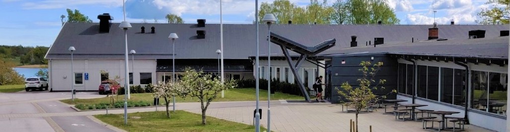 Vit plåtskylt med texten Östra Ringskolan. Skolbyggnader med havet i bakgrunden. 